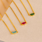 Three gold chain necklaces with rectangular blue, red, and green gemstones are displayed on a textured beige surface, perfect for pairing with your favorite Dainty Marquise Birthstone Necklace.