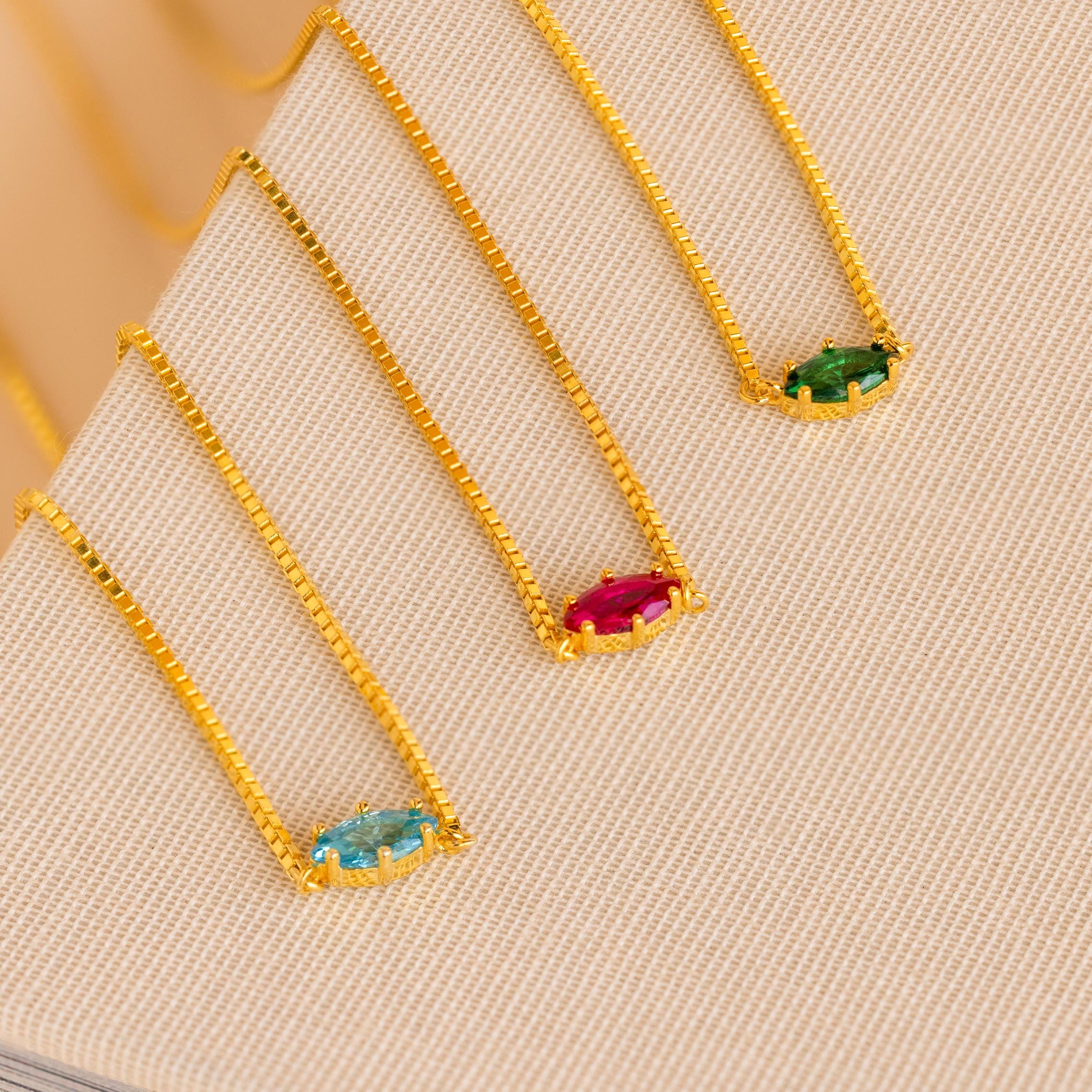 Three gold chain necklaces with rectangular blue, red, and green gemstones are displayed on a textured beige surface, perfect for pairing with your favorite Dainty Marquise Birthstone Necklace.