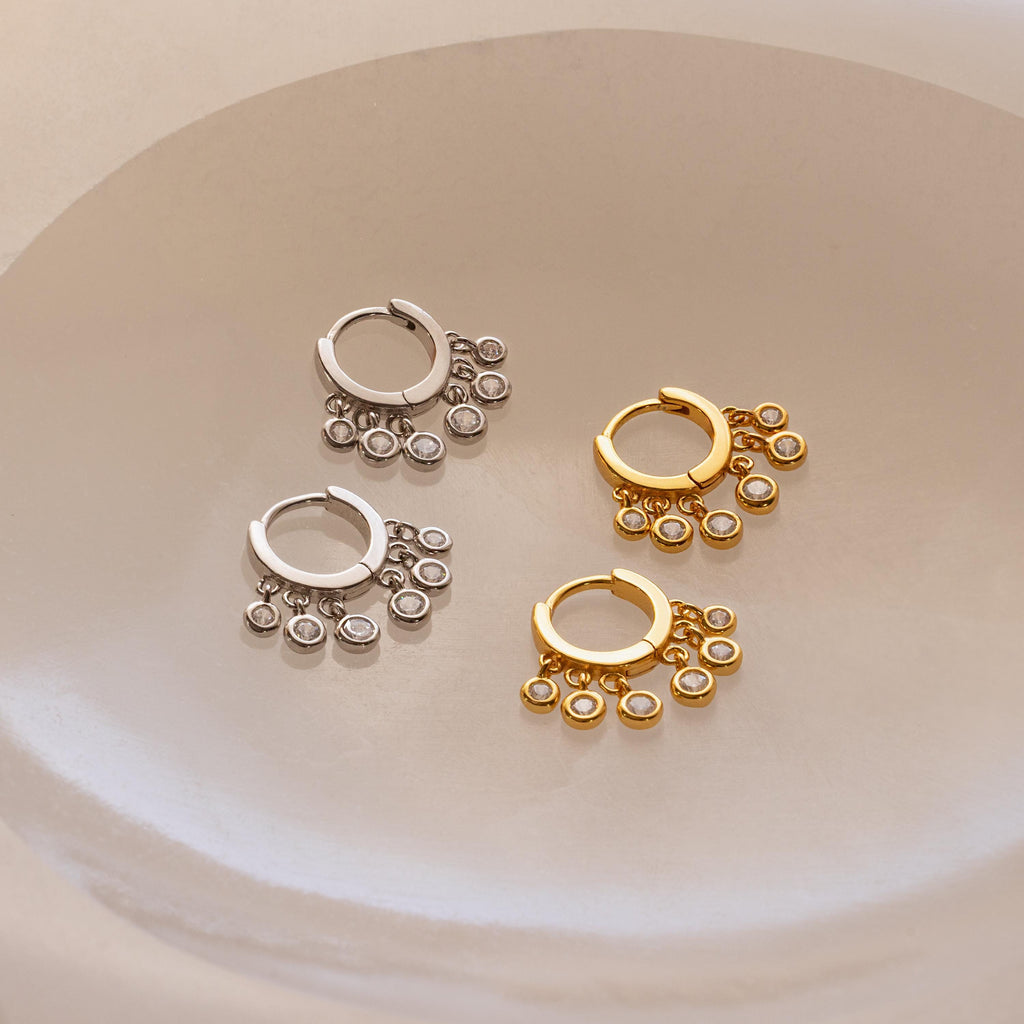 Two pairs of Chandelier Diamond Huggies, featuring an elegant hoop design in silver and gold, are displayed on a smooth, light-colored surface.