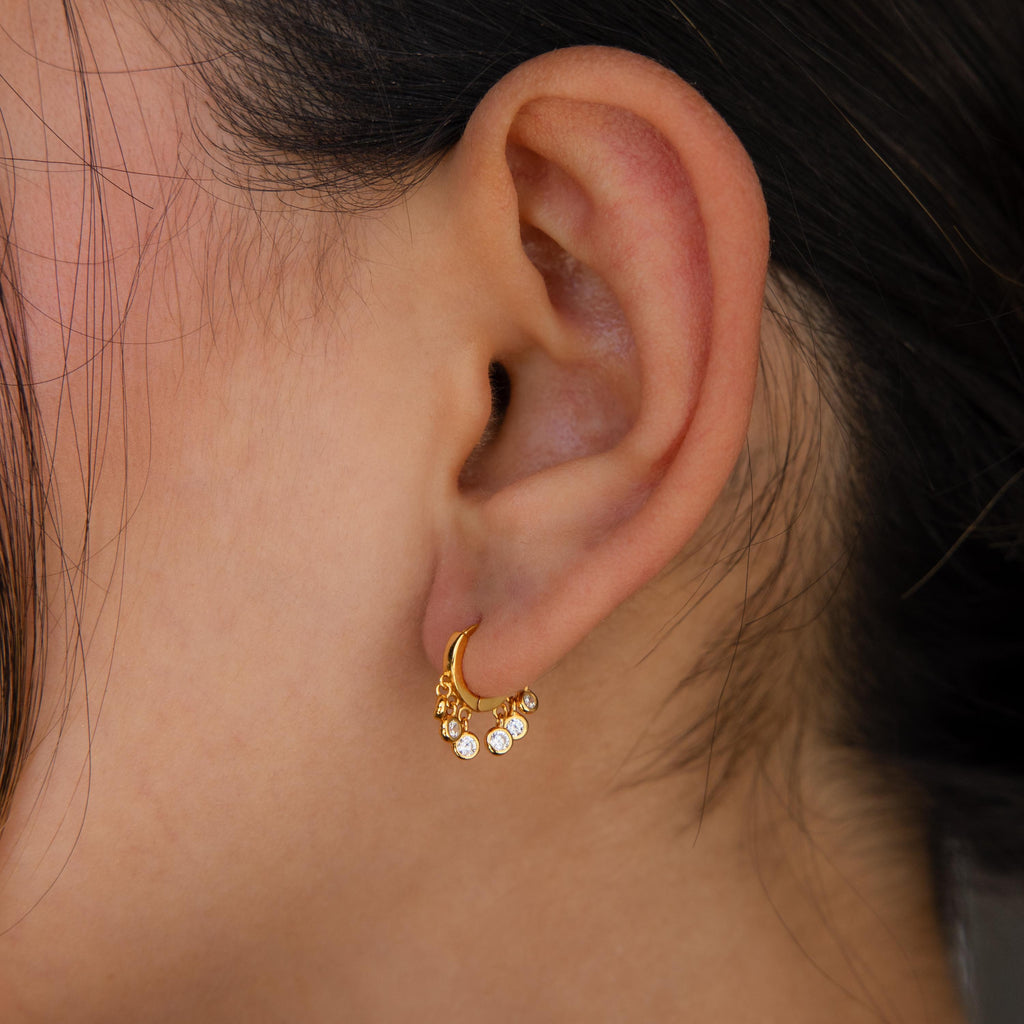 Close-up of a woman's ear wearing Chandelier Diamond Huggies—small gold hoops with dangling crystal diamonds, evoking art deco style.