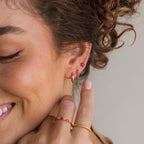 A woman with curly brown hair smiles as she touches her ear, wearing Ivy Flower Birthstone Studs and pink gemstone rings.
