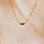 The Dainty Marquise Birthstone Necklace features a gold chain with a small marquise-cut green gemstone pendant, shown on a soft beige fabric background.