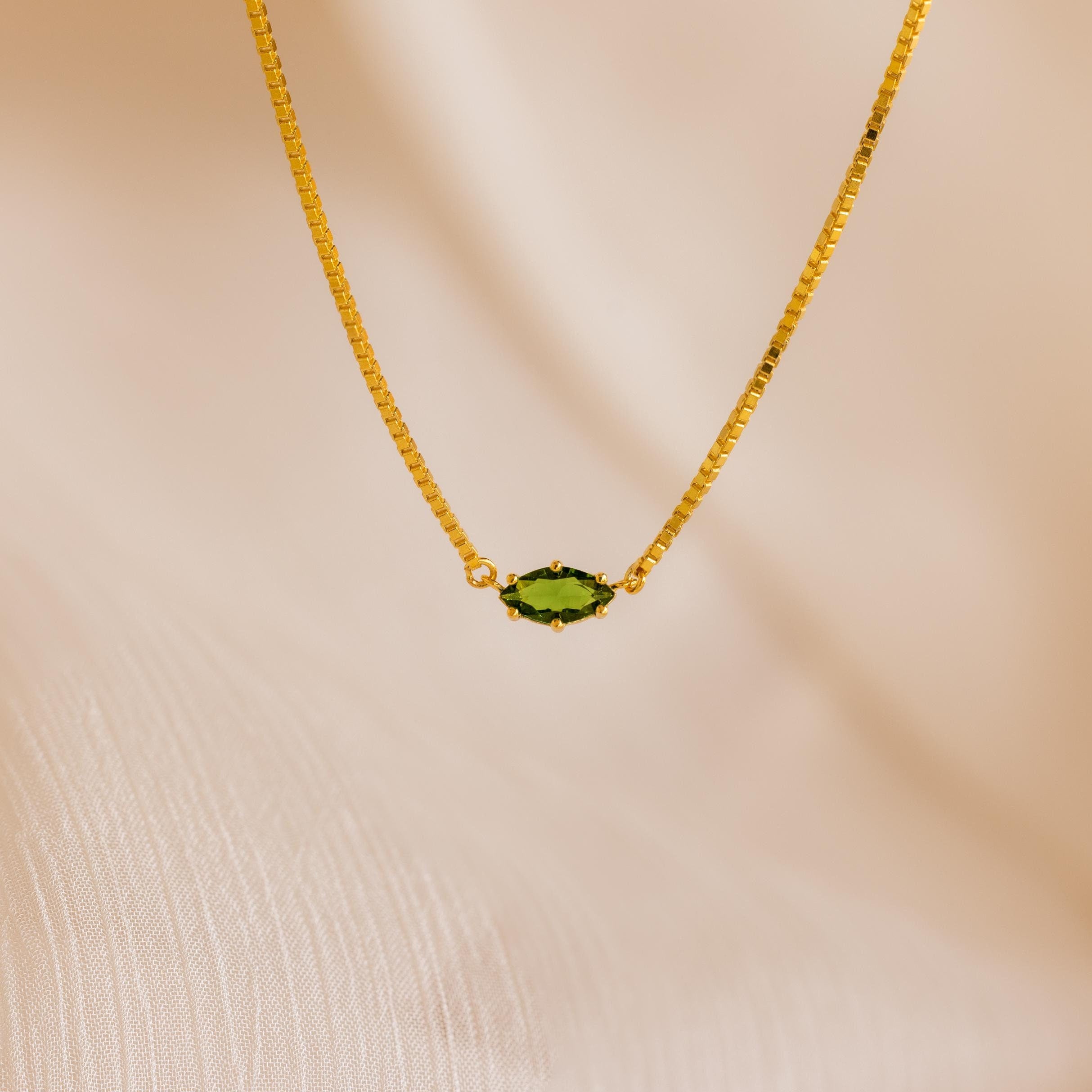 The Dainty Marquise Birthstone Necklace features a gold chain with a small marquise-cut green gemstone pendant, shown on a soft beige fabric background.