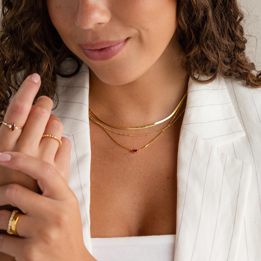 A woman, shown from shoulders to lips, wears a white pinstripe blazer, layered gold necklaces including the Dainty Marquise Birthstone Necklace, and multiple gold rings with red stones.