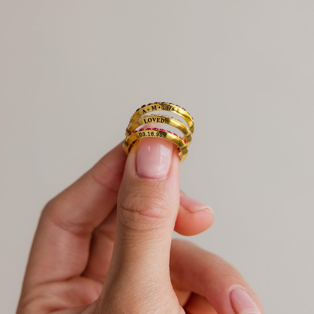 A hand holds four stacked Engraved Birthstone Eternity Rings, each personalized with words and a date, against a plain background.
