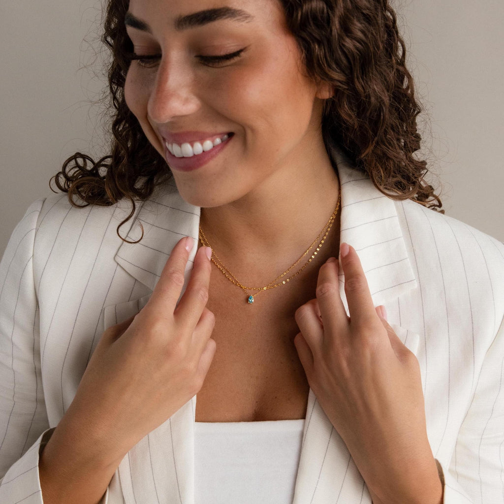 A woman in a white pinstripe blazer smiles while looking down, her hands gently adjusting the Duo Layered Birthstone Necklace, which features a gold design with a pear-cut birthstone and small pendant.