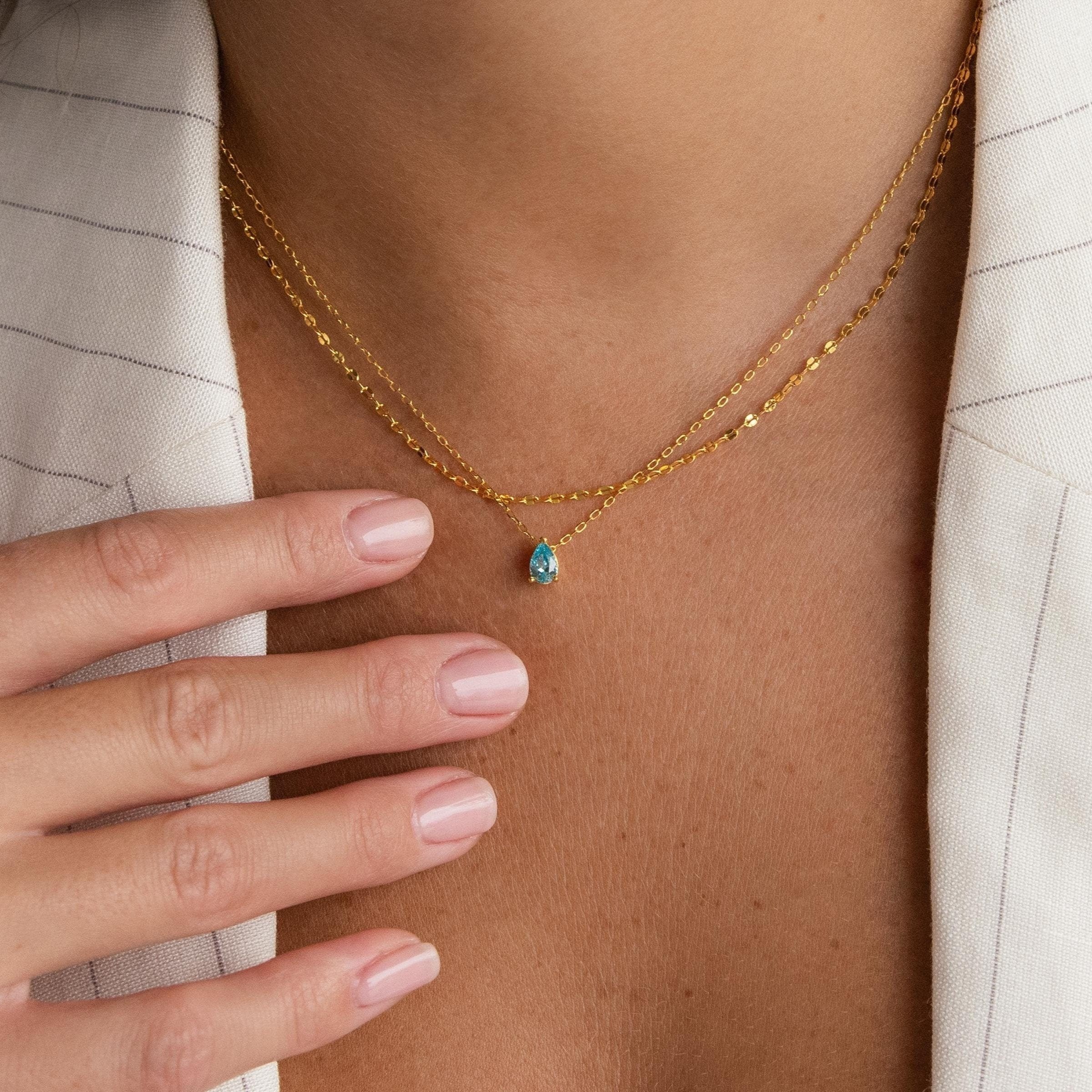 A person wears a beige pinstripe blazer and the Duo Layered Birthstone Necklace, featuring a blue pear-cut pendant in sterling silver, with neatly manicured nails resting near their collarbone.