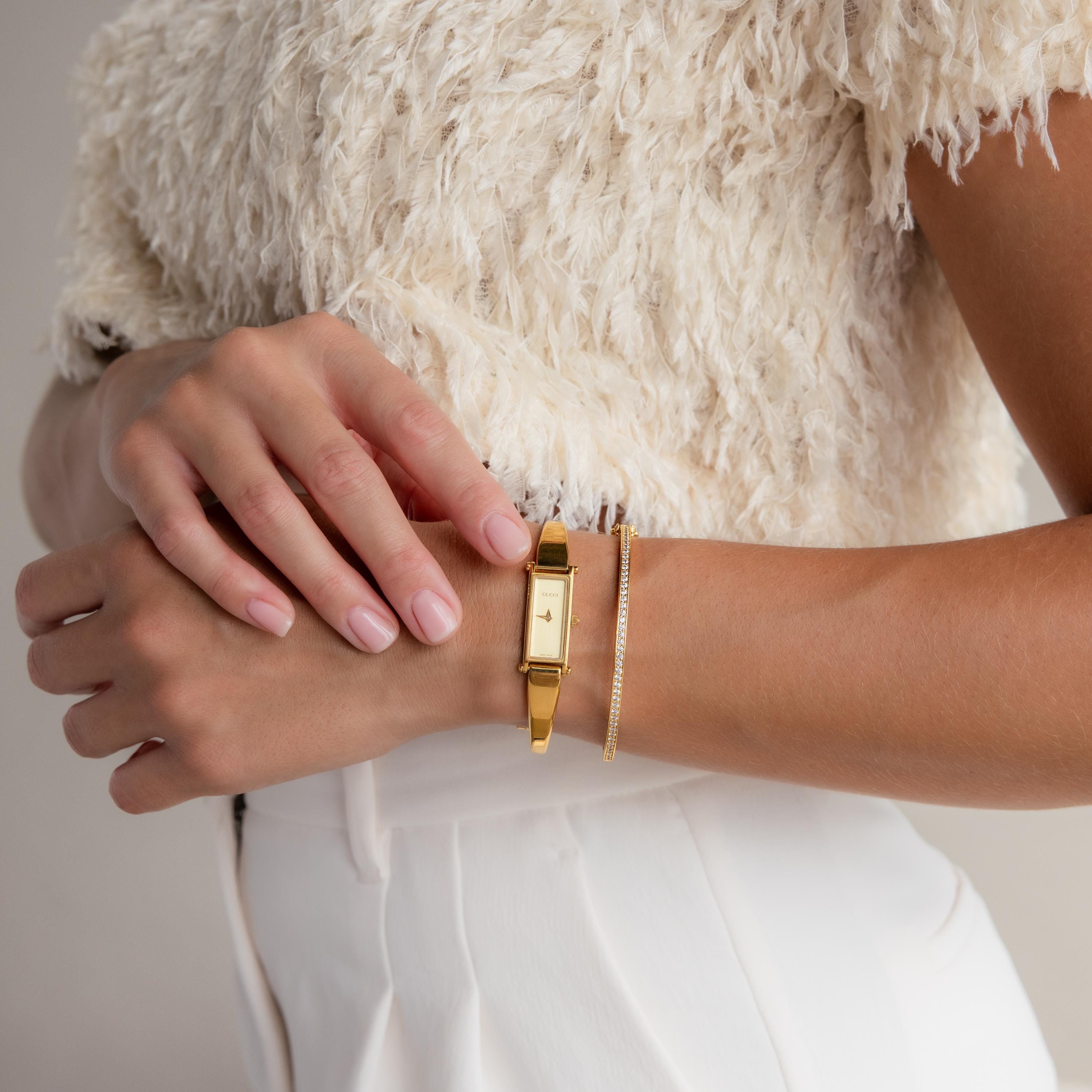 Woman wearing a Diamond Tennis Bangle featuring radiant diamonds, paired with a gold watch, styled in a white textured top and white pants.