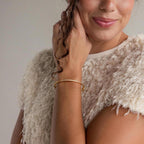 A woman touches her curled hair and smiles softly, wearing a textured cream top and the Diamond Tennis Bangle on her wrist.