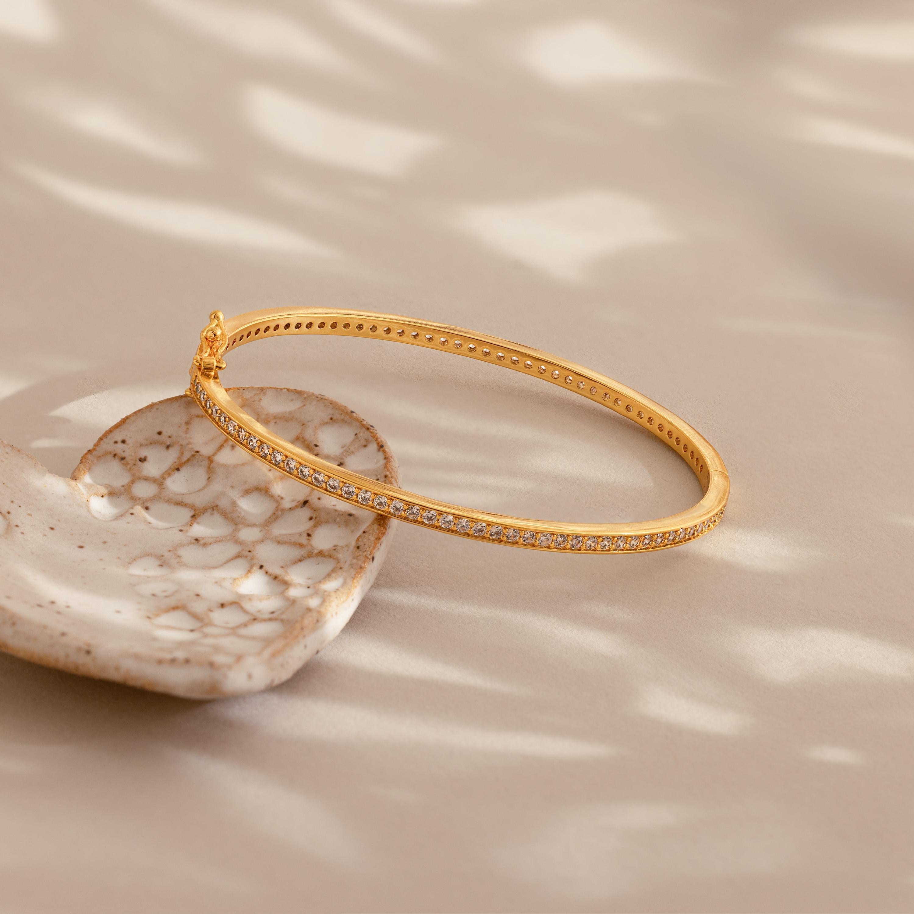 The Diamond Tennis Bangle, featuring radiant diamonds and a curved design, rests on a textured ceramic dish and catches the light in soft shadows.