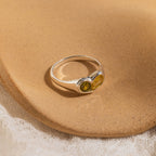 The Duo Birthstone Heart Signet Ring, featuring two yellow-green gemstones, is displayed on a textured beige surface.