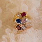 The Duo Birthstone Heart Signet Ring, a heart-shaped gemstone ring, is stacked with others in gold, silver, and rose gold atop cream-colored lace fabric.