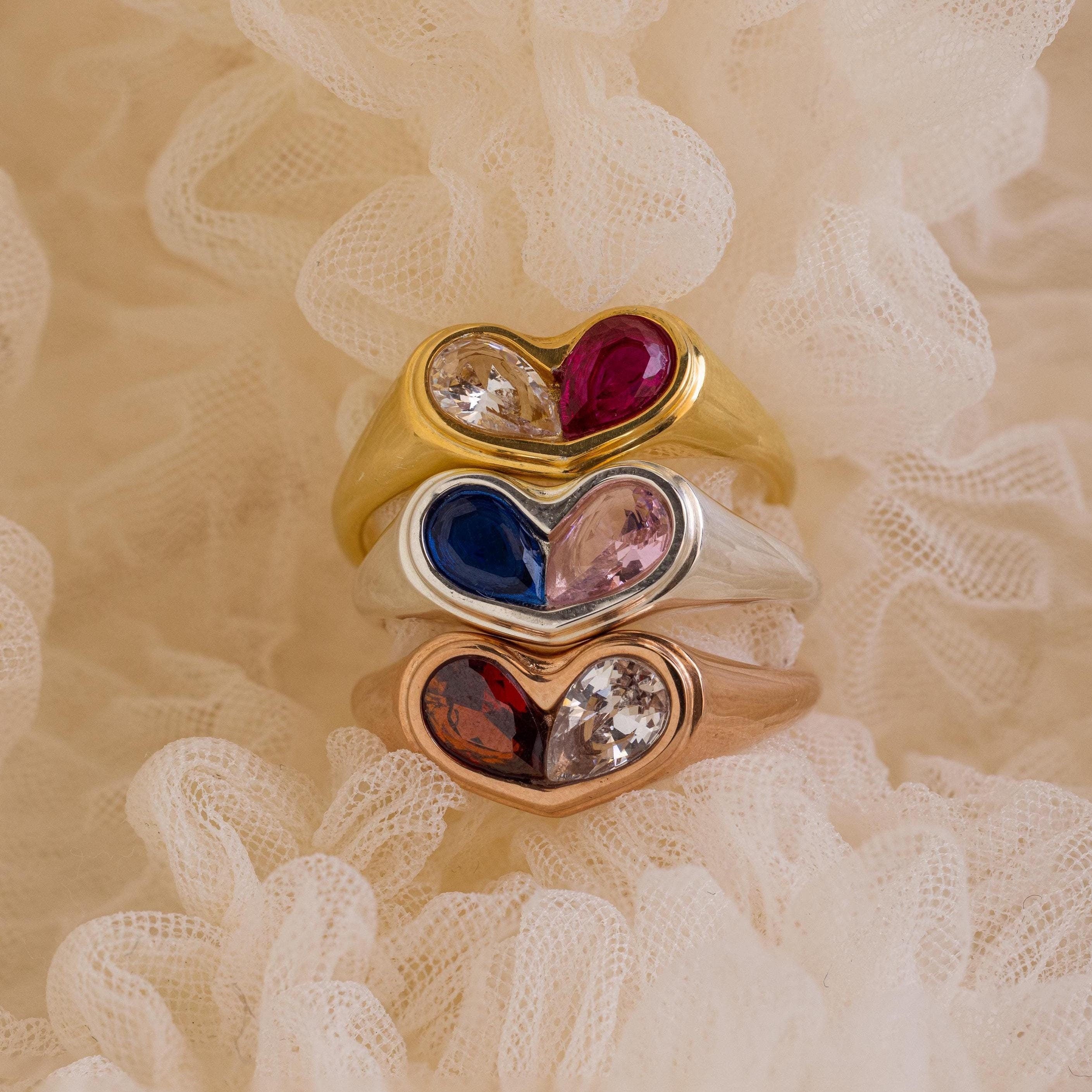 The Duo Birthstone Heart Signet Ring, a heart-shaped gemstone ring, is stacked with others in gold, silver, and rose gold atop cream-colored lace fabric.