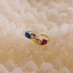 The Duo Birthstone Heart Signet Ring, crafted in gold and adorned with blue, clear, and red gemstones, rests on soft cream-colored fabric.