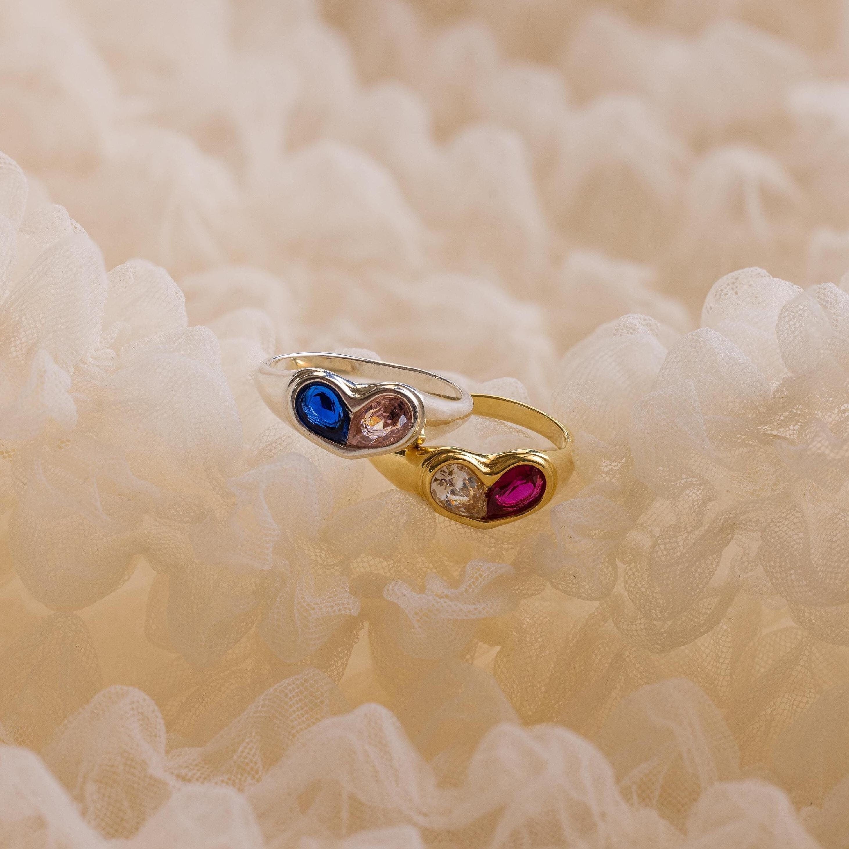 The Duo Birthstone Heart Signet Ring, crafted in gold and adorned with blue, clear, and red gemstones, rests on soft cream-colored fabric.