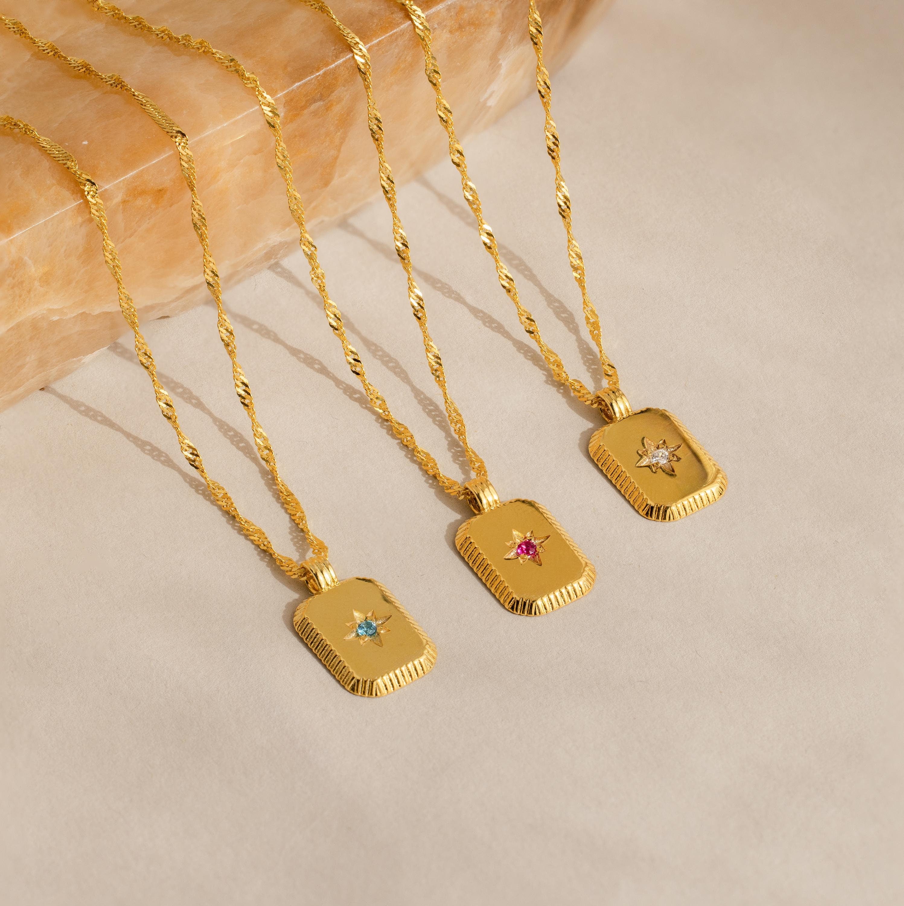 The Ribbed Birthstone Star Necklace, a gold necklace with a rectangular pendant featuring a unique gemstone, is displayed on stone. Its ribbed design and personalized birthstone make it both stylish and meaningful.
