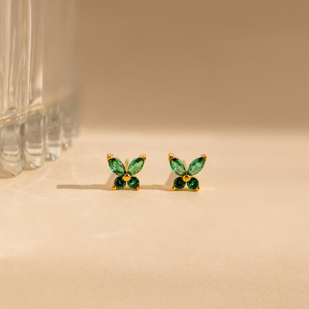 The Butterfly Birthstone Studs feature green, leaf-shaped stones set in gold, reminiscent of butterfly jewelry, and are showcased on a beige surface next to a glass.
