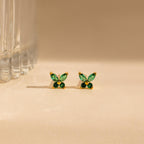 The Butterfly Birthstone Studs feature green, leaf-shaped stones set in gold, reminiscent of butterfly jewelry, and are showcased on a beige surface next to a glass.
