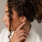 A woman with curly hair, dressed in a white blazer, touches a Butterfly Birthstone Studs earring on her left ear.