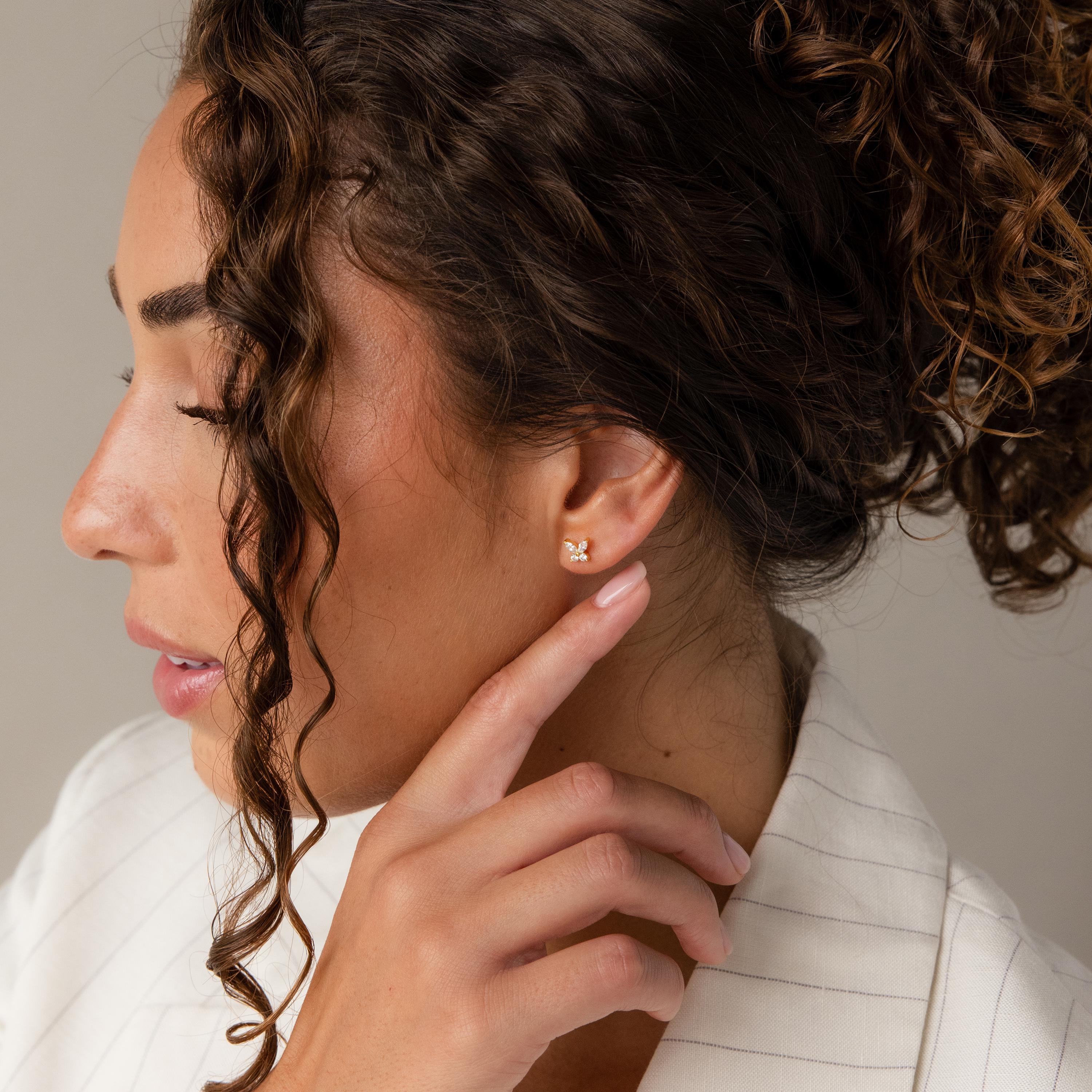A woman with curly hair, dressed in a white blazer, touches a Butterfly Birthstone Studs earring on her left ear.
