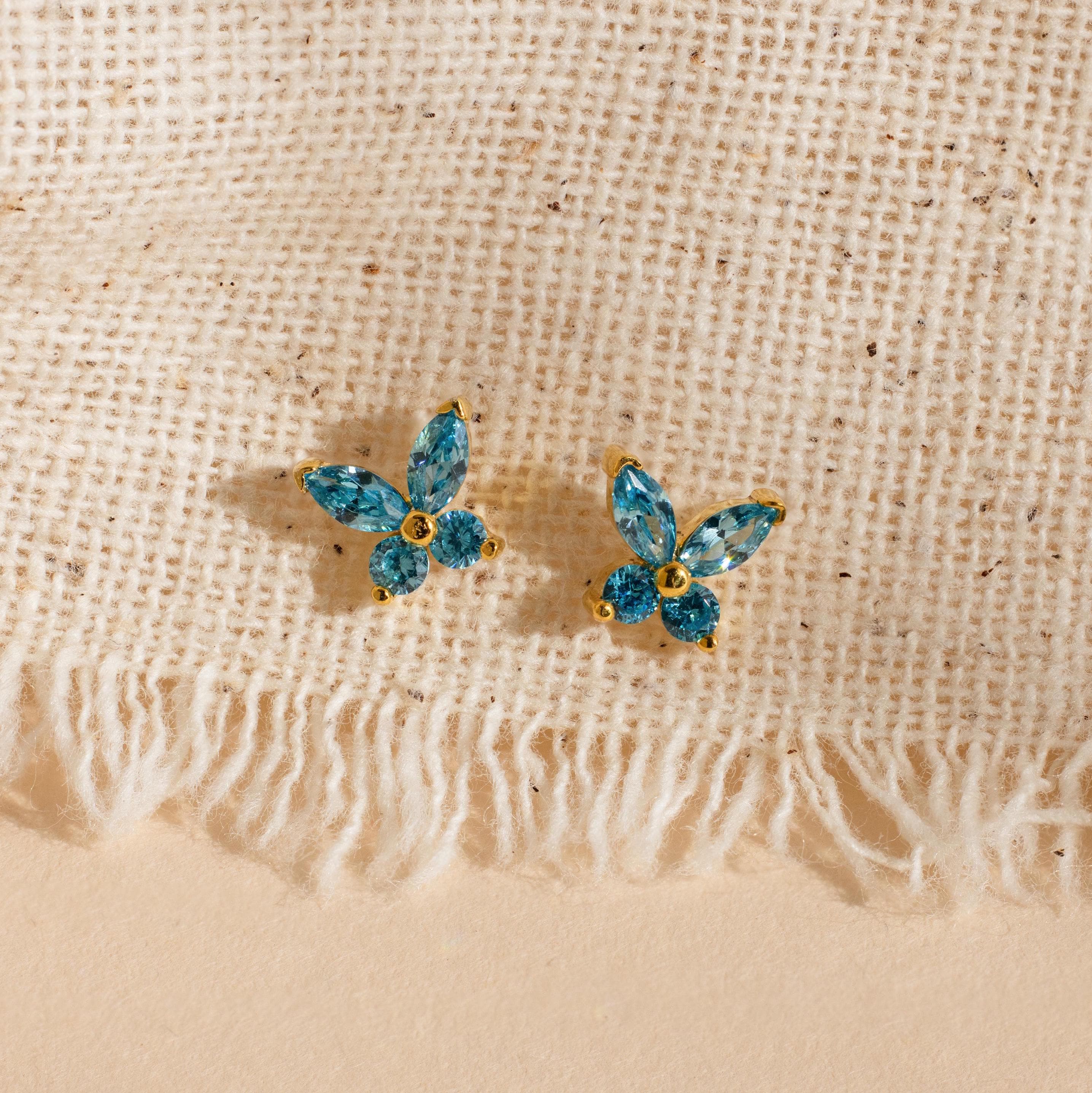 Butterfly Birthstone Studs with blue gemstones are displayed on a textured beige fabric background.