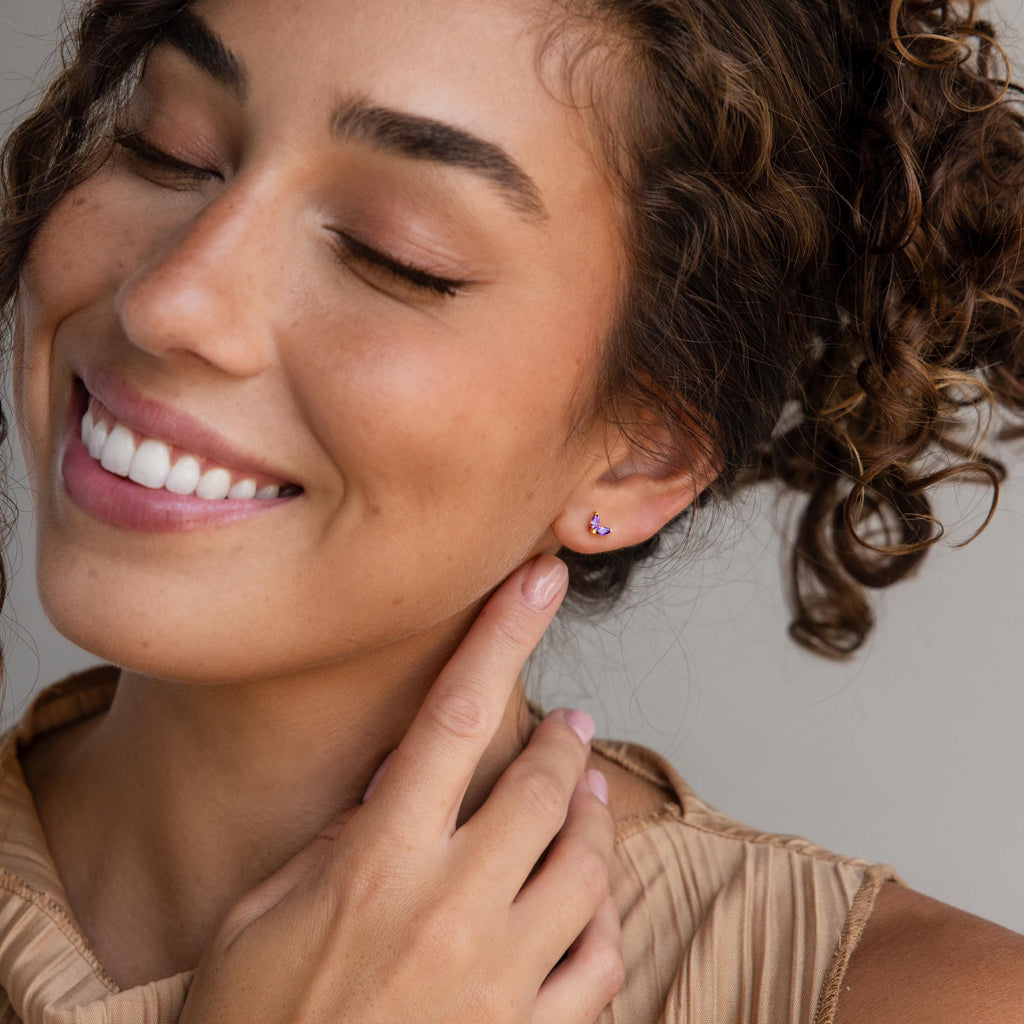 A smiling woman with curly hair touches her ear, showing off a Marquise Petal Birthstone Stud in Amethyst (February)—an elegant choice for lovers of personalized jewelry.