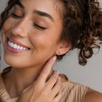 A smiling woman with curly hair touches her ear, showing off a Marquise Petal Birthstone Stud in Amethyst (February)—an elegant choice for lovers of personalized jewelry.