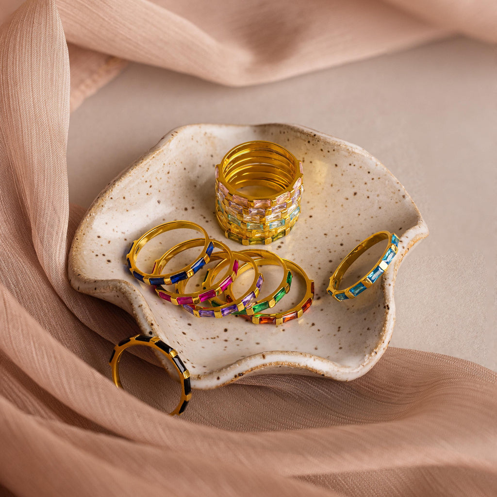 The Half Eternity Birthstone Ring, a gold ring set with colorful gemstones, is displayed on a speckled ceramic dish and surrounded by soft beige fabric.