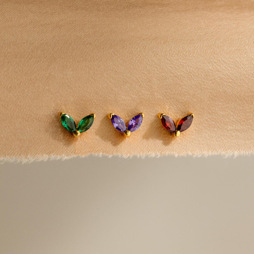 Three pairs of Marquise Petal Birthstone Studs—ideal as birthstone earrings or personalized jewelry—sparkle in green, purple, and red tones on a beige fabric background.