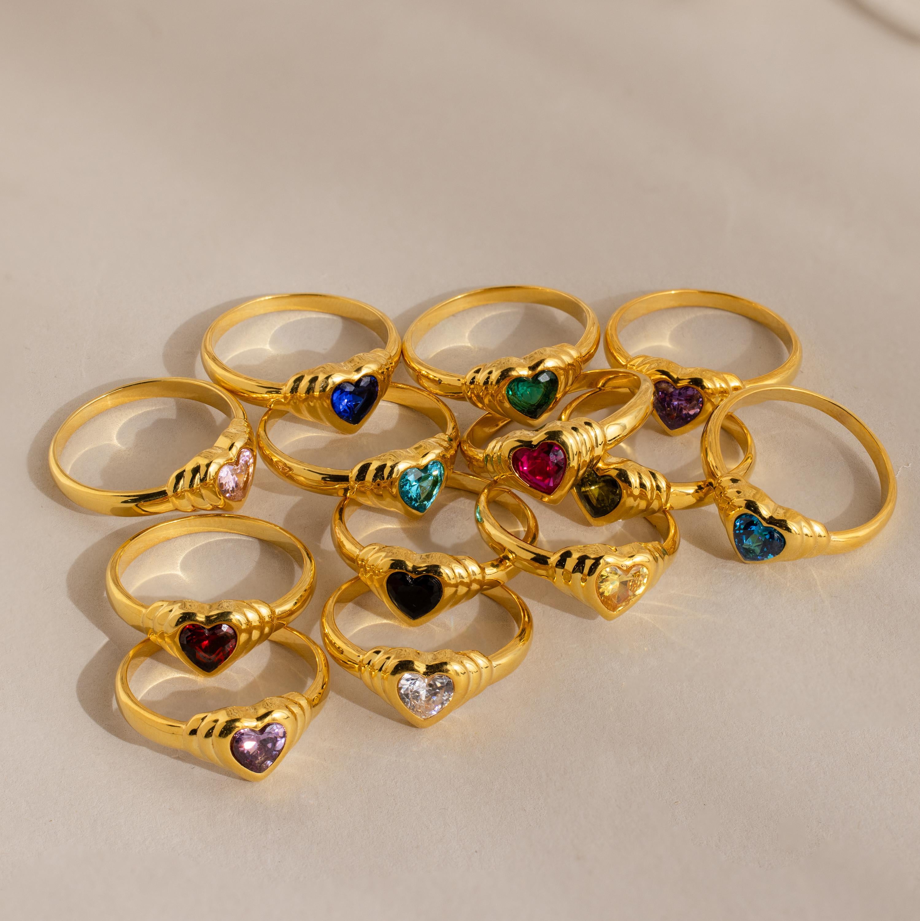 Twelve Birthstone Heart Signet Rings, each featuring a heart-shaped gemstone in a unique color, are displayed on a beige surface.