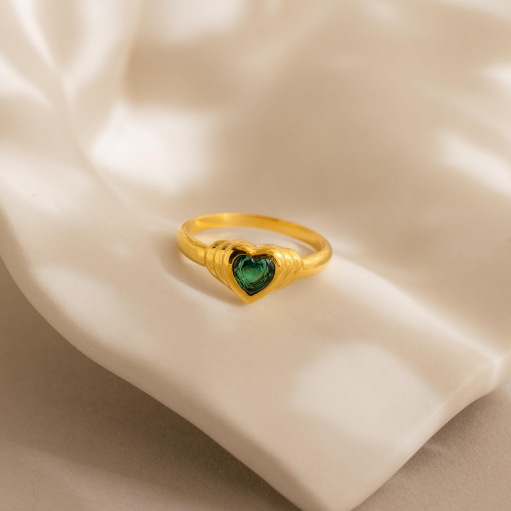 The Birthstone Heart Signet Ring showcases a green heart-shaped gemstone, beautifully presented on cream-colored fabric.