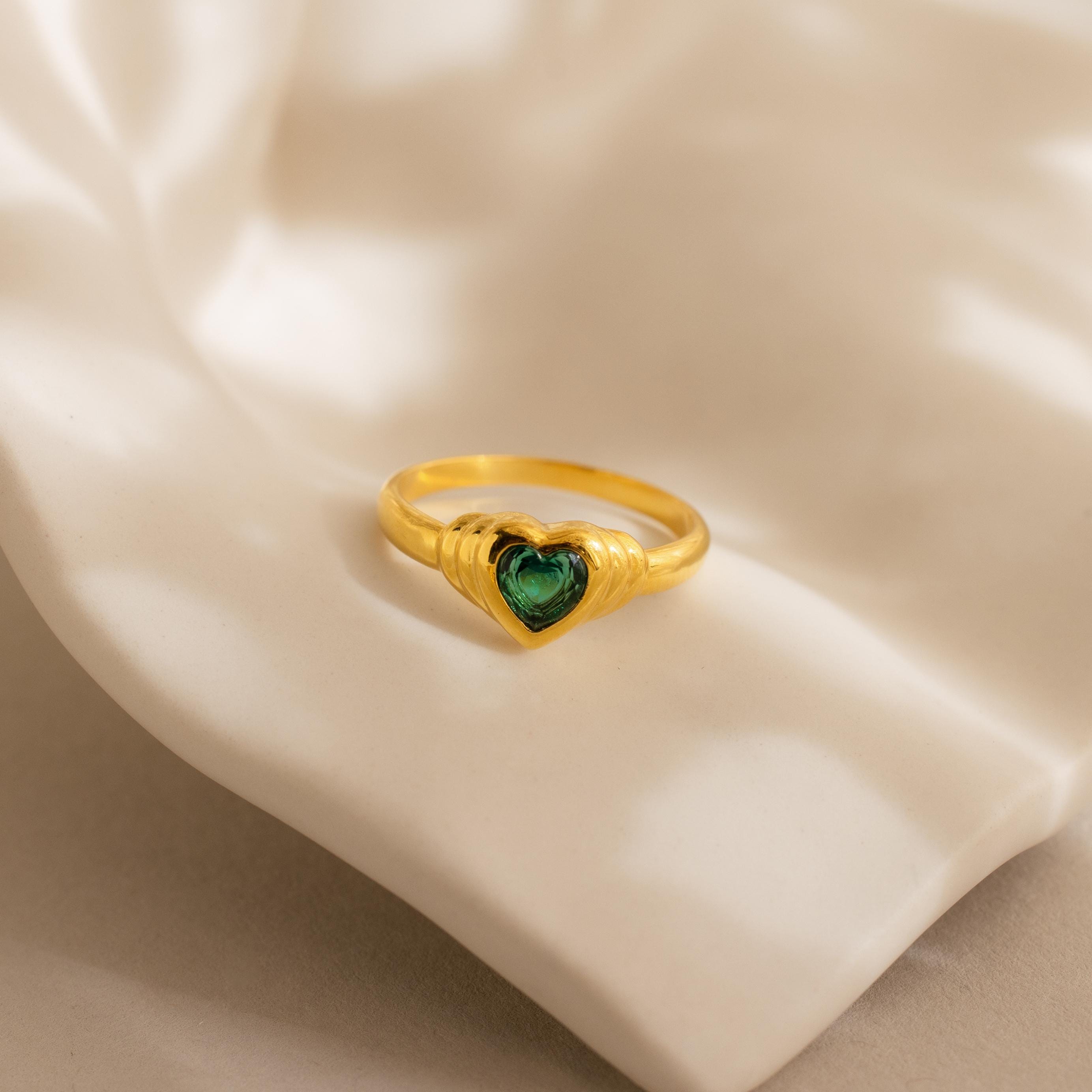 The Birthstone Heart Signet Ring showcases a green heart-shaped gemstone, beautifully presented on cream-colored fabric.