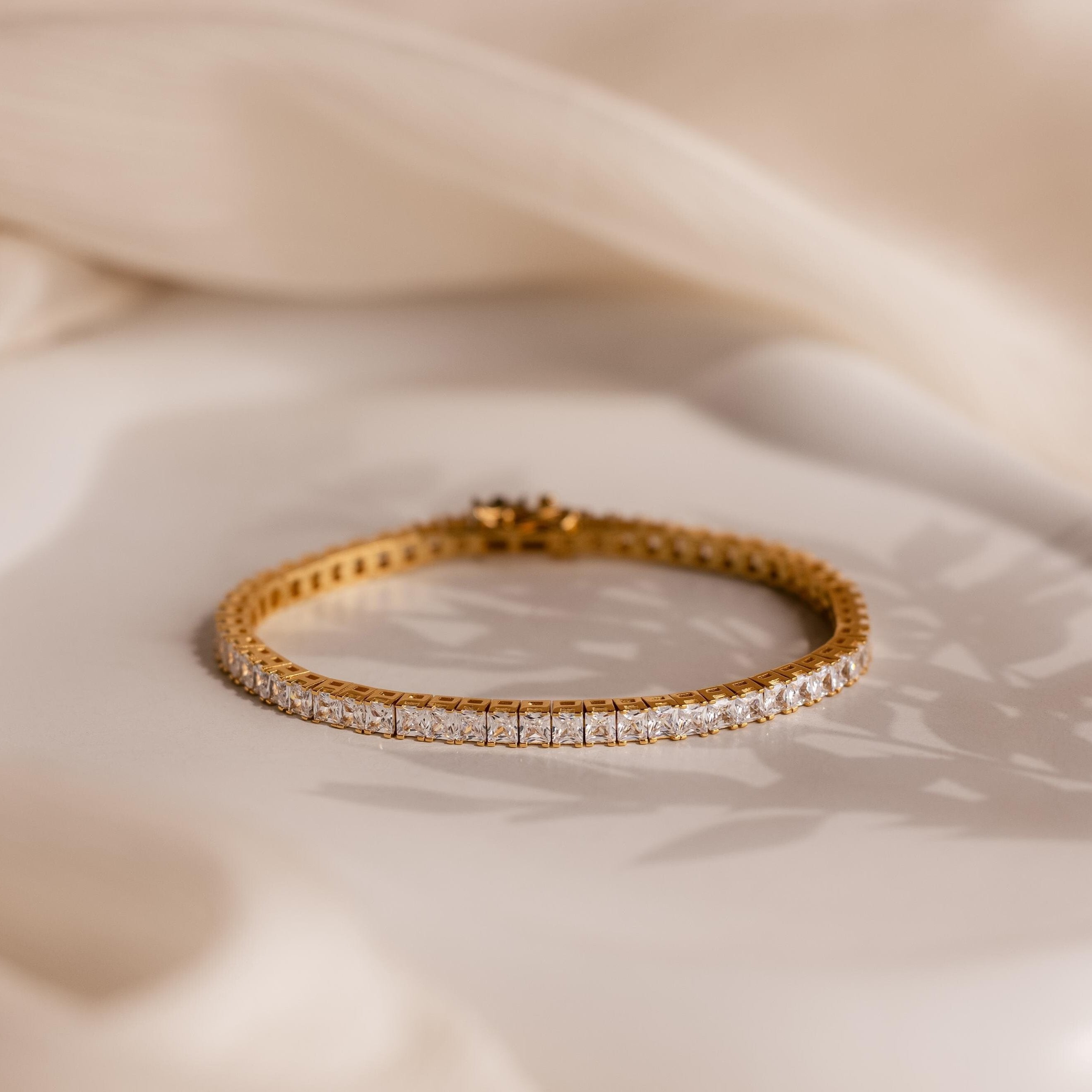 The Princess Diamond Tennis Bracelet features clear, princess-cut diamonds displayed on a soft beige fabric background with subtle shadows.