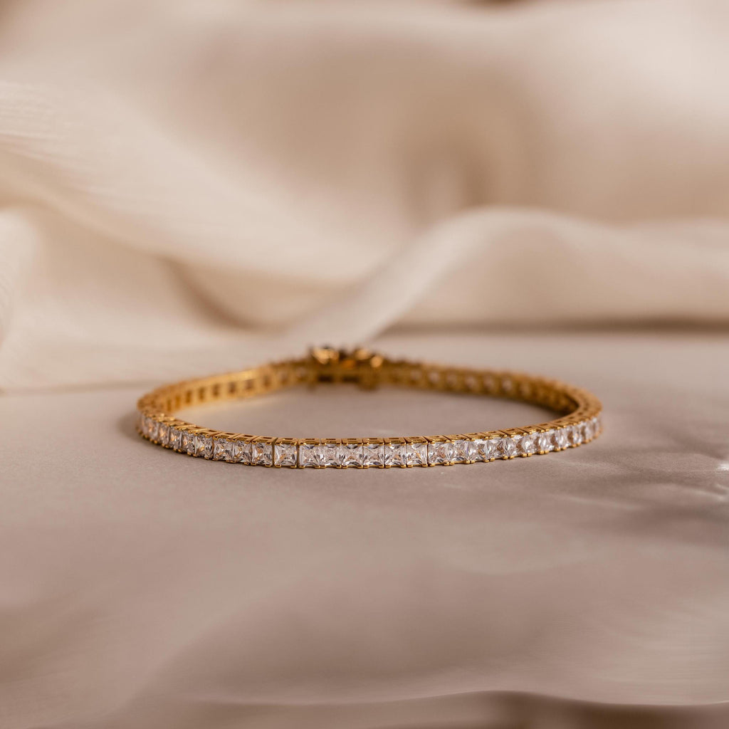 The Princess Diamond Tennis Bracelet in 18K Gold features sparkling princess-cut diamonds, elegantly showcased on a soft beige fabric background.