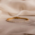 The Princess Diamond Tennis Bracelet in 18K Gold features sparkling princess-cut diamonds, elegantly showcased on a soft beige fabric background.