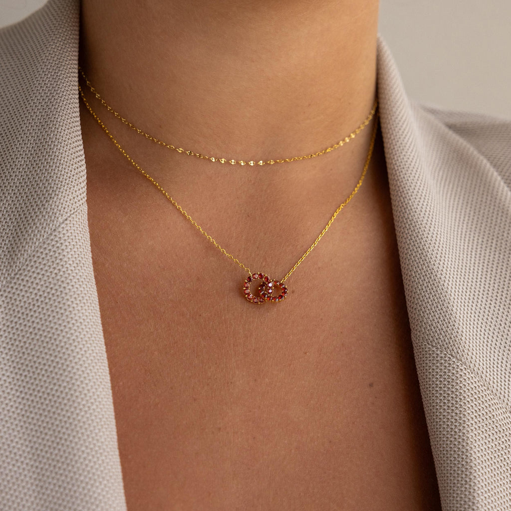Woman wearing a beige blazer and two gold necklaces: one with a red heart-shaped pendant, the other the Interlocking Duo Birthstone Necklace.
