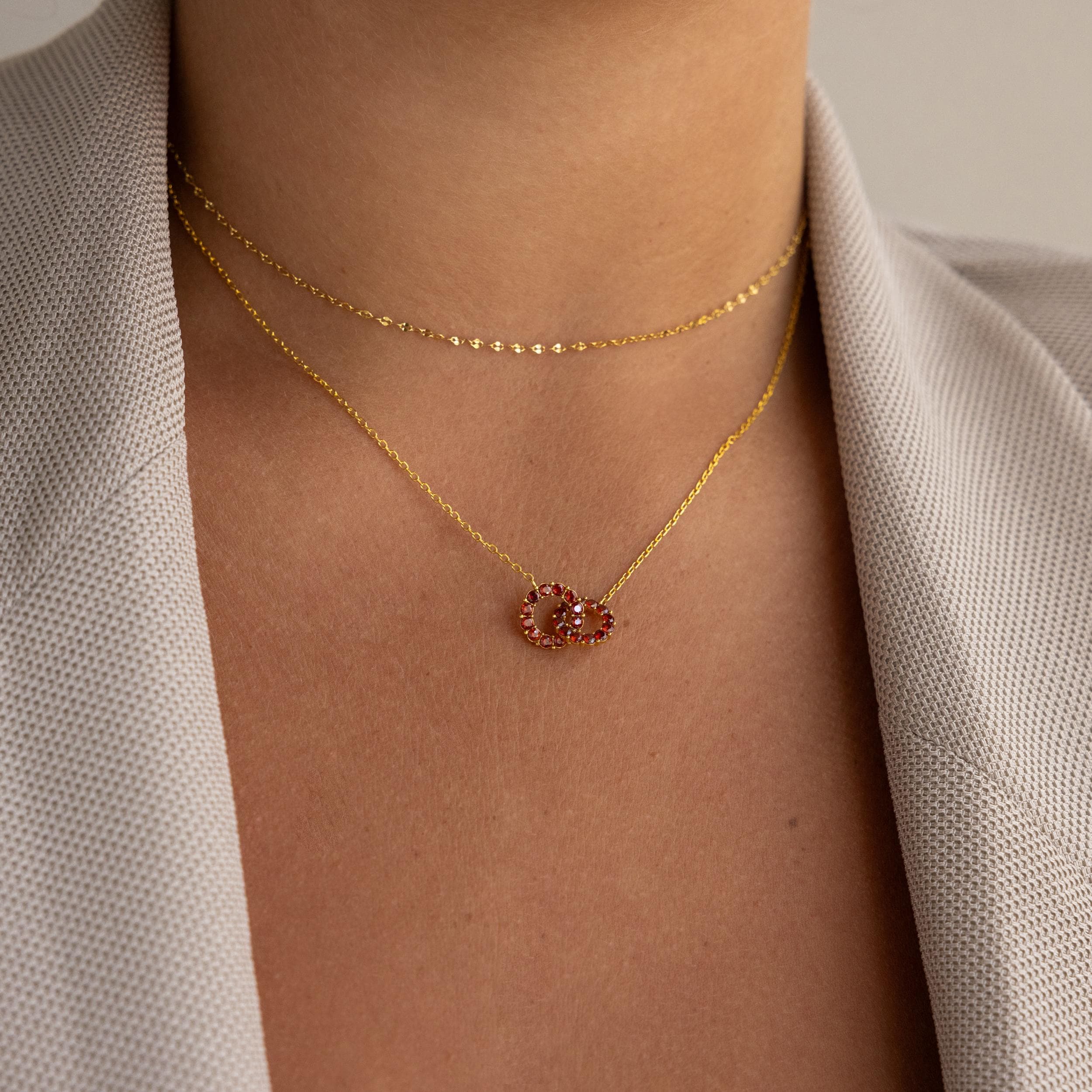 Woman wearing a beige blazer and two gold necklaces: one with a red heart-shaped pendant, the other the Interlocking Duo Birthstone Necklace.