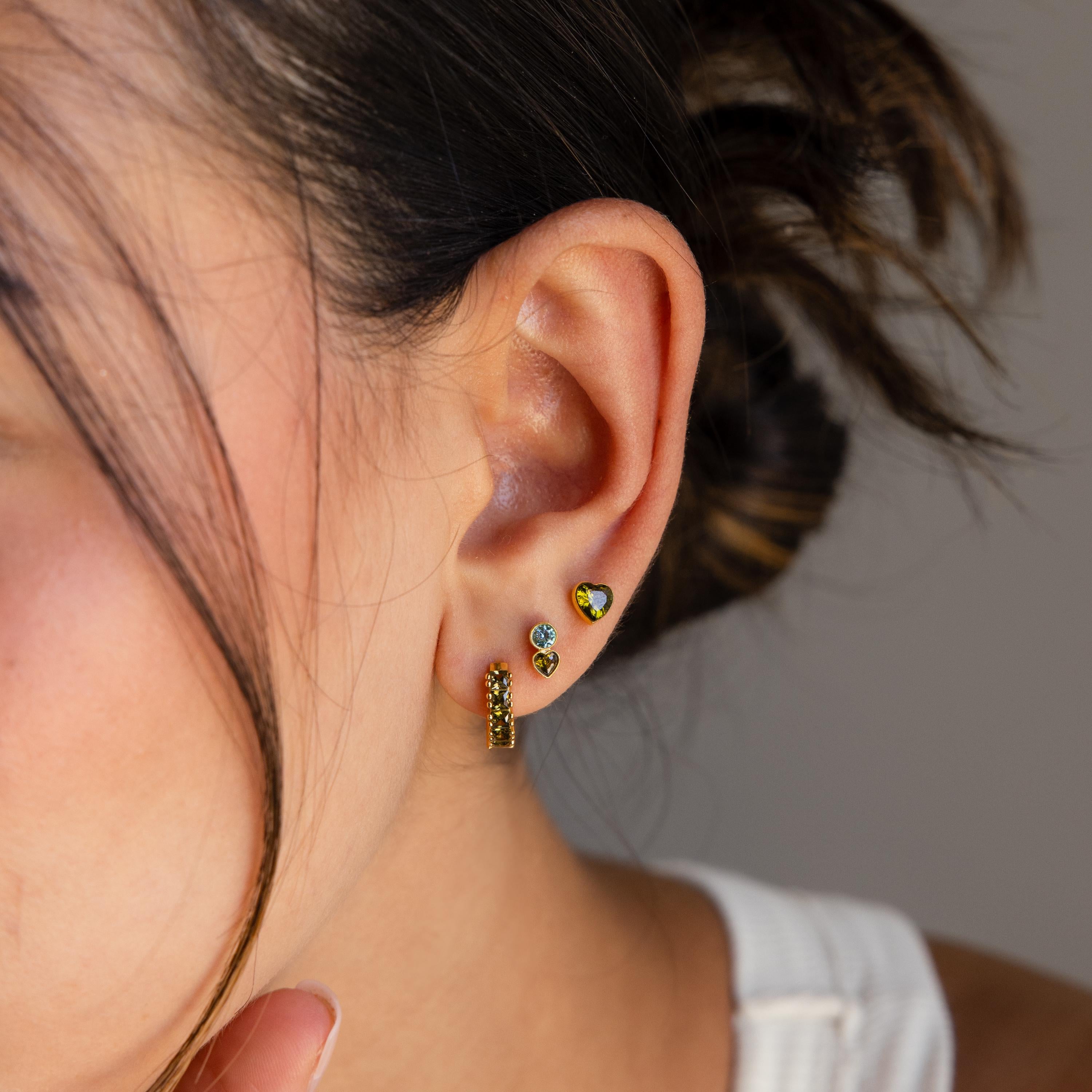 A woman's ear adorned with three gold and gemstone earrings, including the Toi et Moi Birthstone Studs. With her hair tied back and a white top, these studs are shown as an ideal personalized earring gift.