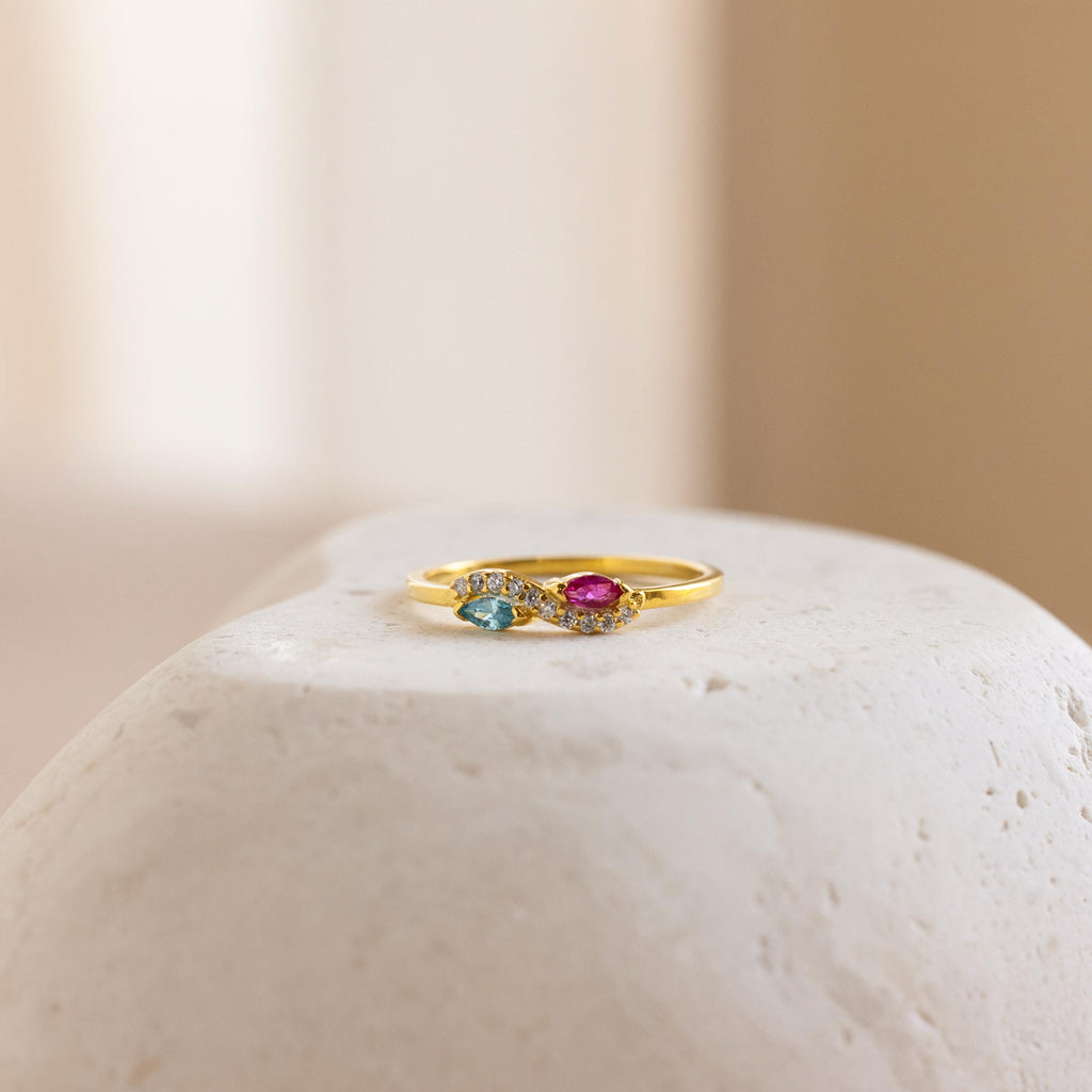 Infinity Marquise Birthstone Ring, featuring blue and pink gemstones, is elegantly displayed on a smooth white stone with a soft, neutral background.