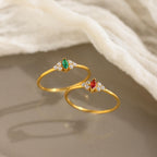 The Tiny Marquise Birthstone Ring, featuring a delicate gold band with a marquise-cut gemstone and small diamonds, is pictured with two rings—one green stone and one red—on a soft backdrop. A unique, customizable piece for elegant style.