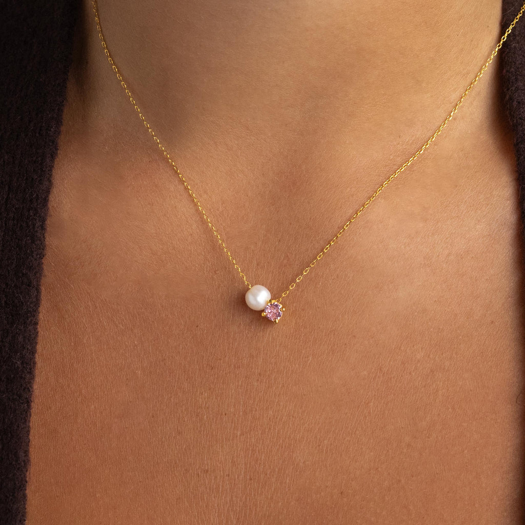 The Pearl & Birthstone Necklace is a personalized gold jewelry piece featuring a delicate pink birthstone pendant and elegant pearl accents, designed to rest gracefully on her neck.