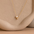 The Pearl & Birthstone Necklace features a gold chain with a single pearl and small gemstone pendant, ideal as personalized or birthstone jewelry, shown on a textured beige surface.