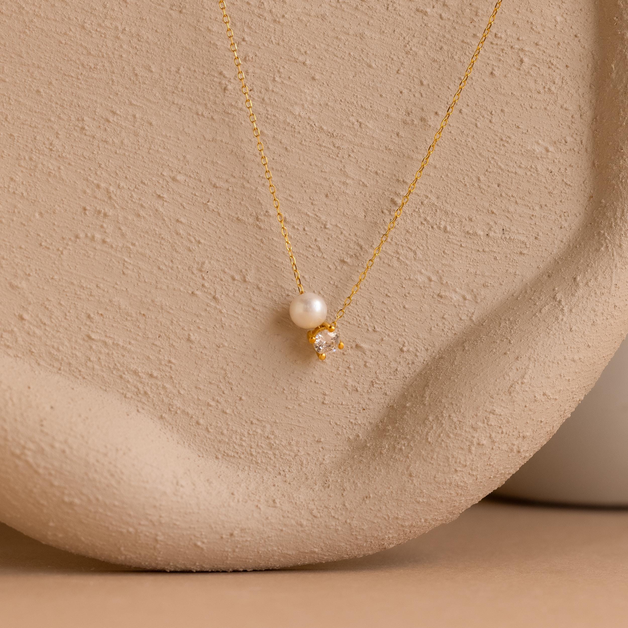 The Pearl & Birthstone Necklace features a gold chain with a single pearl and small gemstone pendant, ideal as personalized or birthstone jewelry, shown on a textured beige surface.