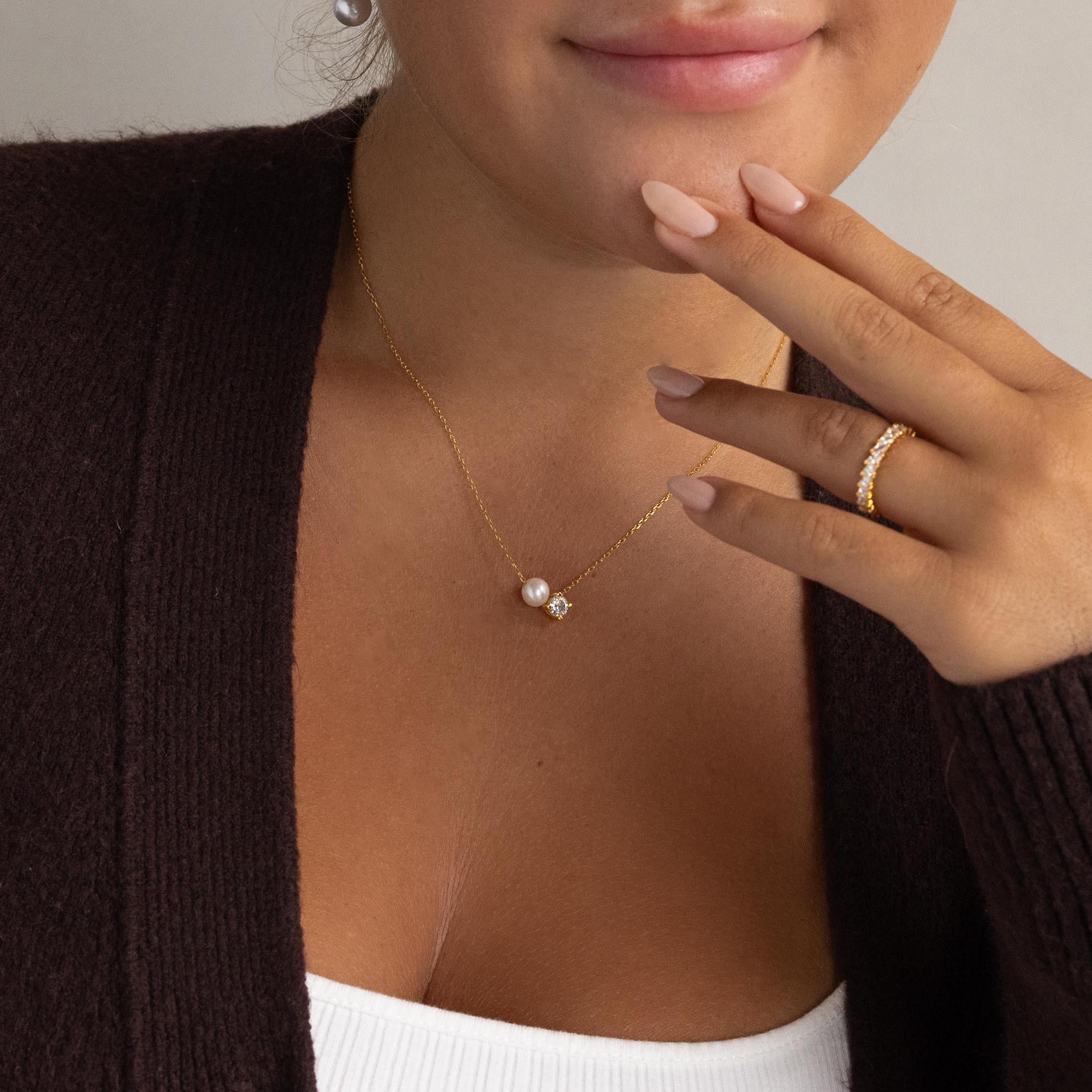 A woman wears the Pearl & Birthstone Necklace, a gold ring, brown cardigan, and white top—an elegant showcase of personalized jewelry that’s perfect for any occasion.