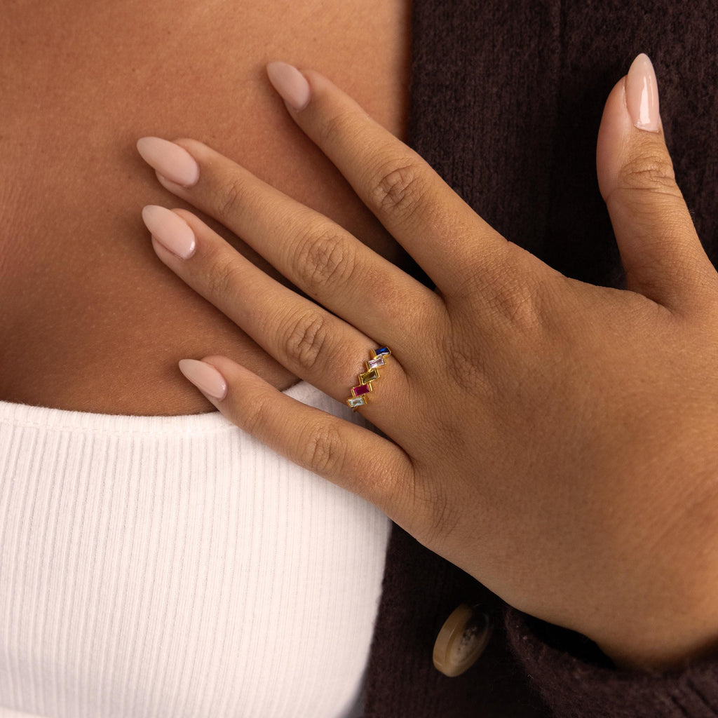A manicured hand wearing the Multiple Baguette Birthstone Ring rests on a chest, styled with a white top and brown cardigan.