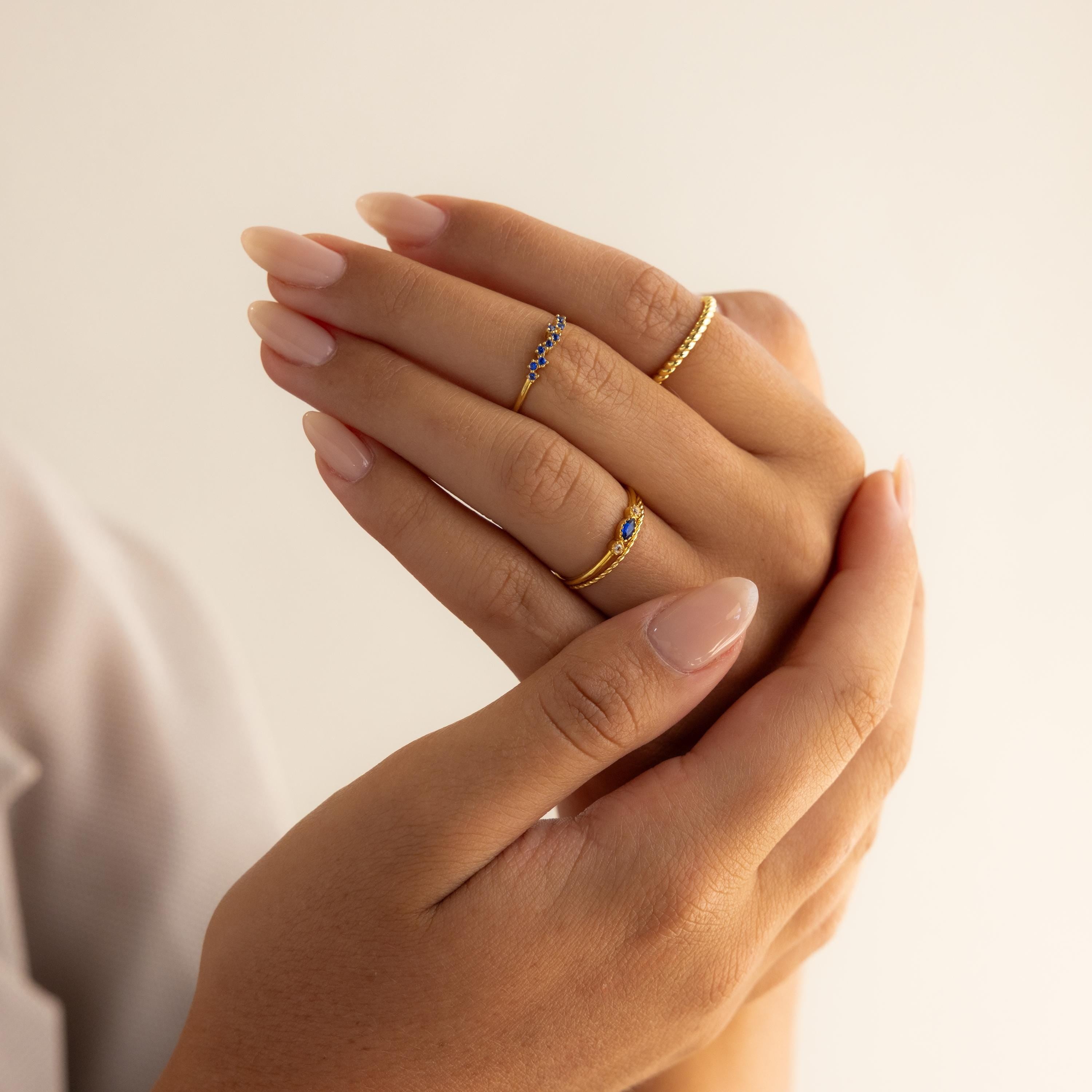 Manicured hands wear delicate gold rings, featuring the Cluster Marquise Birthstone Ring and dainty blue marquise-cut bands, all set against a neutral background.