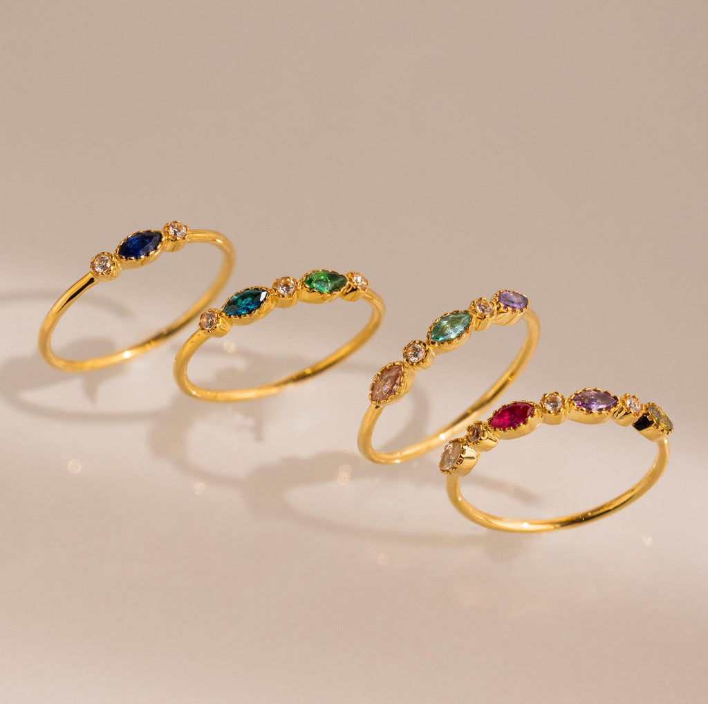 A Cluster Marquise Birthstone Ring is displayed among four gold rings with colorful gemstones and diamonds, all arranged diagonally on a neutral background.