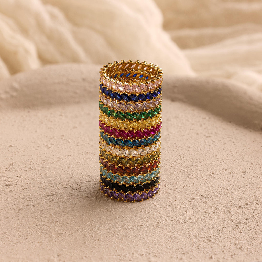 The Eternity Marquise Birthstone Ring, featuring a customizable gold band and colorful gemstones, stands upright on a textured beige surface.