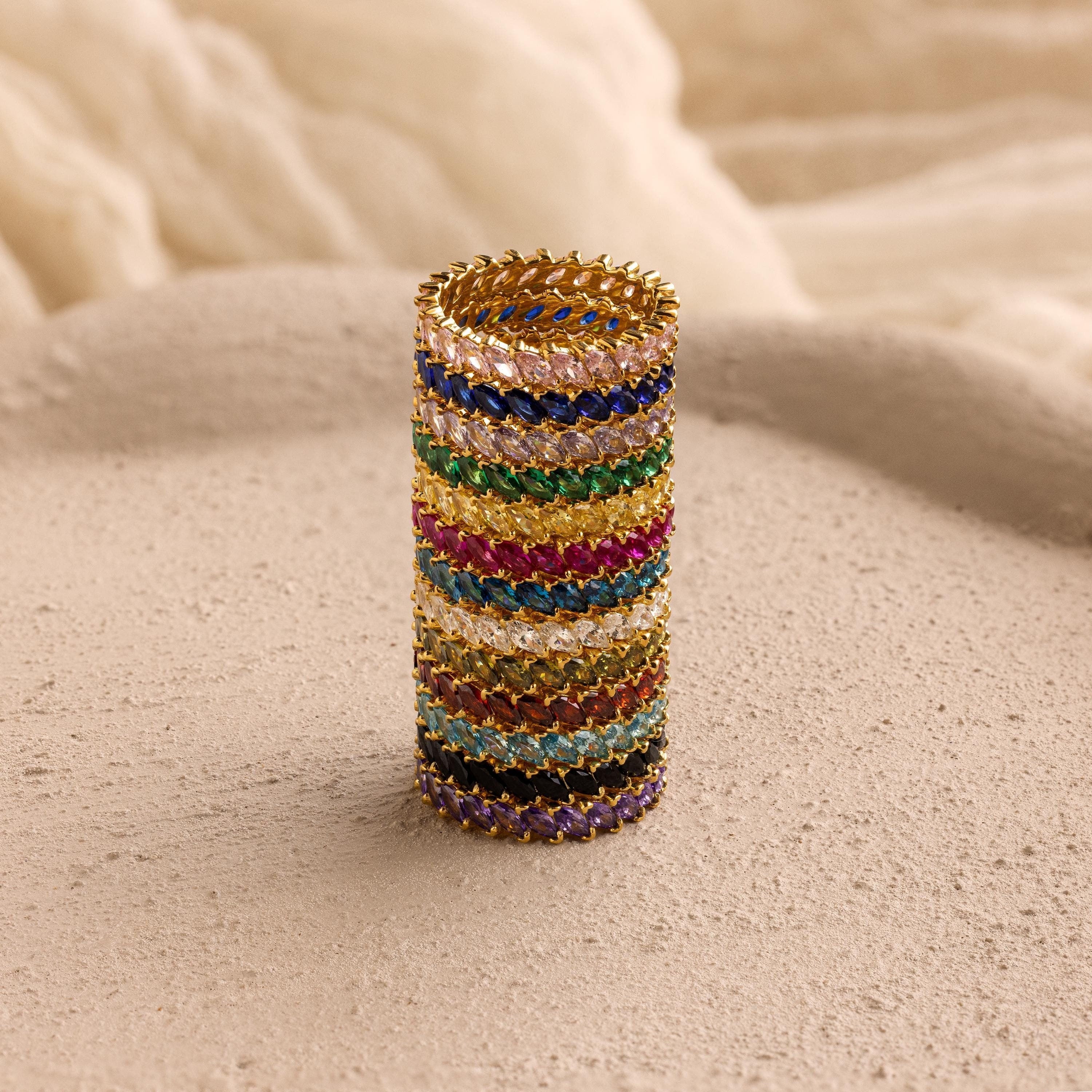 The Eternity Marquise Birthstone Ring, featuring a customizable gold band and colorful gemstones, stands upright on a textured beige surface.