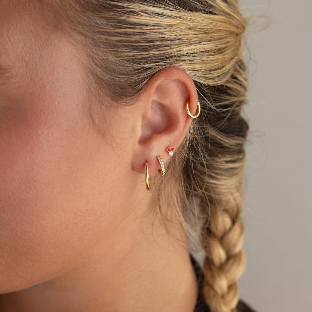 Close-up of a woman's ear adorned with multiple gold earrings, featuring Diamond Marquise Birthstone Studs and a small braid.